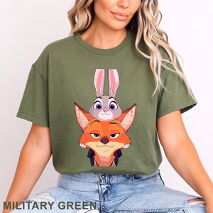 May include: Military green t-shirt featuring a graphic of Judy Hopps and Nick Wilde from Zootopia. The design shows the characters stacked on top of each other. The text "MILITARY GREEN" is at the bottom of the image.