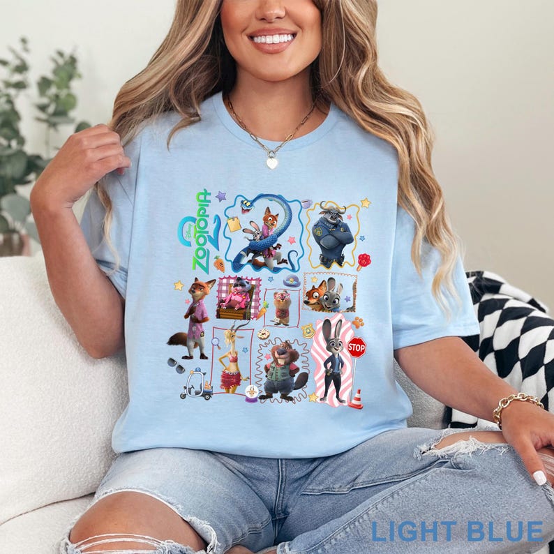 May include: Light blue t-shirt featuring a colorful graphic of characters from the animated film Zootopia. The design includes various animal characters in a grid layout, with the film's title in green. The shirt also has the text "LIGHT BLUE" at the bottom.