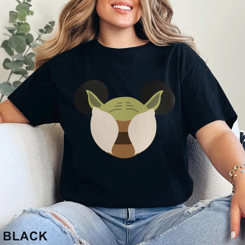 May include: Black t-shirt featuring a graphic of a green character with large ears, resembling a popular movie character. The character's face is designed with white, brown, and green elements. The word "BLACK" is printed at the bottom.