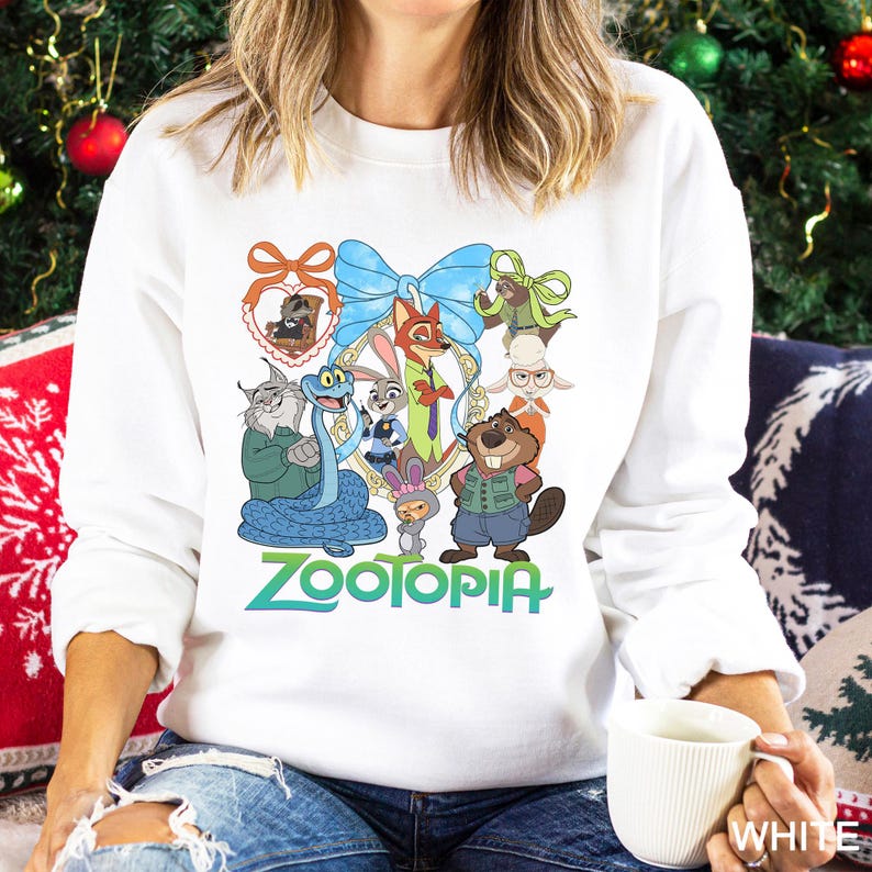 May include: White crew neck sweatshirt with a colourful graphic of characters from the animated film "Zootopia." The design includes a blue bow, a gold frame, and the word "Zootopia" in green and blue. The sweatshirt is a crew neck style.
