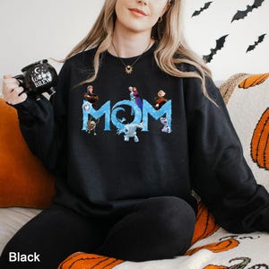 K&ouml;nnte beinhalten: Ein schwarzes Langarm-Sweatshirt mit dem Wort MOM in gro&szlig;en, eisblauen Buchstaben. Das Design enth&auml;lt verschiedene animierte Figuren aus dem Film Die Eisk&ouml;nigin, die in den Text integriert sind. Eine schwarze Tasse mit dem Text Witches Brew.