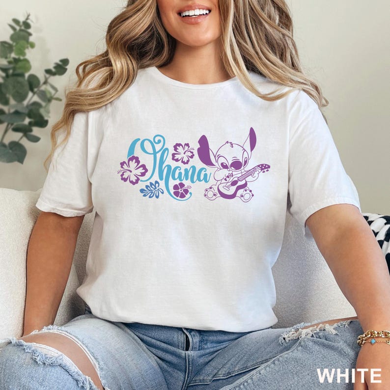 Op de afbeelding: Een wit T-shirt met korte mouwen en een paarse afbeelding van het personage Stitch die ukelele speelt, vergezeld door de blauwe tekst "Ohana" en paarse illustraties van hibiscusbloemen. Het ontwerp staat centraal op de borst.