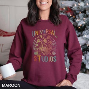 Pode incluir: Camisola bord&ocirc; com gola redonda com um gr&aacute;fico colorido de personagens da Universal Studios e o texto "UNIVERSAL STUDIOS". O design inclui um castelo, Minions e outras imagens ic&oacute;nicas. A camisola &eacute; de cor bord&ocirc; lisa.