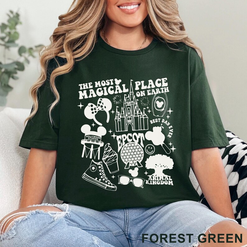 May include: Forest green t-shirt with a white graphic design featuring the text "The Most Magical Place on Earth" and various Disney-themed icons, including a castle, Mickey Mouse ears, and a smiling face.
