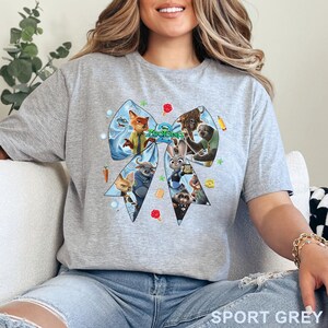 Op de afbeelding: Sportgrijs t-shirt met een groot, lichtblauw strikdesign met personages uit de film "Zootopia". Het strikdesign bevat afbeeldingen van een vos, een konijn en andere dierenpersonages. De tekst "Zootopia" staat in het midden van de strik.