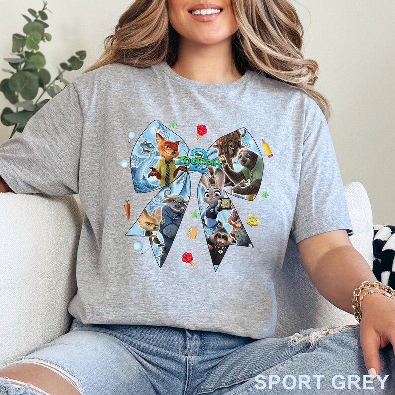 Op de afbeelding: Sportgrijs t-shirt met een groot, lichtblauw strikdesign met personages uit de film "Zootopia". Het strikdesign bevat afbeeldingen van een vos, een konijn en andere dierenpersonages. De tekst "Zootopia" staat in het midden van de strik.