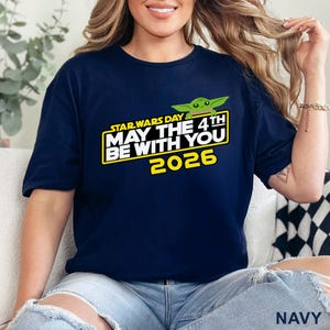 May include: Navy blue t-shirt featuring a graphic with the text "STAR WARS DAY MAY THE 4TH BE WITH YOU 2026" and a green cartoon character. The shirt is a short-sleeved crew neck.