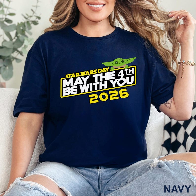 May include: Navy blue t-shirt featuring a graphic with the text "STAR WARS DAY MAY THE 4TH BE WITH YOU 2026" and a green cartoon character. The shirt is a short-sleeved crew neck.