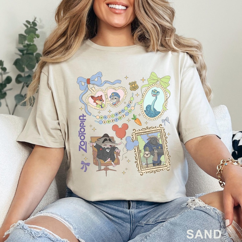 May include: Sand-colored t-shirt featuring colorful cartoon illustrations from the movie Zootopia. The design includes framed portraits of characters, a heart-shaped frame with Nick and Judy, and the word "Zootopia" vertically on the left side.
