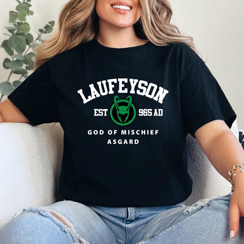 May include: Black t-shirt with the text "LAUFEYSON" above a green and white graphic. Below the graphic are the words "EST 965 AD GOD OF MISCHIEF ASGARD". The shirt is a crew neck.