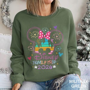 May include: Olive green sweatshirt with a colorful Disney graphic. The design includes a Minnie Mouse bow, fireworks, and the words "Disney Family Trip 2026". The sweatshirt is a comfortable, casual style.
