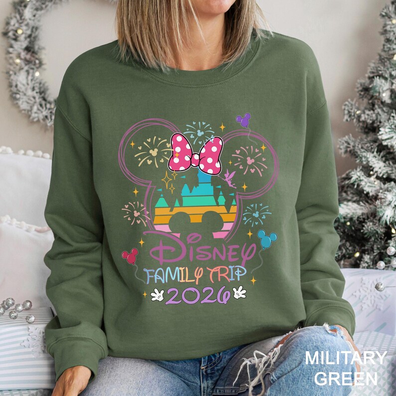 May include: Olive green sweatshirt with a colorful Disney graphic. The design includes a Minnie Mouse bow, fireworks, and the words "Disney Family Trip 2026". The sweatshirt is a comfortable, casual style.