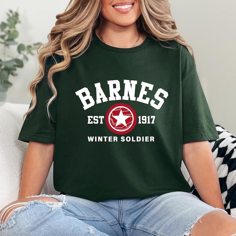 May include: Dark green t-shirt featuring the word "BARNES" in white block letters above a red circle with a white star. Below the circle are the words "EST 1917" and "WINTER SOLDIER" in white. The shirt is made of a soft material.