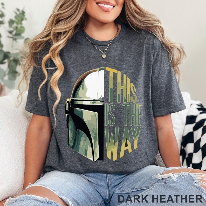 Pu&ograve; includere: Una t-shirt a maniche corte grigio melange scuro con una stampa grafica di un casco metallico sul lato sinistro e il testo THIS IS THE WAY in lettere maiuscole gialle e verdi sul lato destro, con un motivo mimetico sovrapposto.