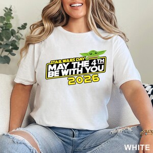 May include: White t-shirt featuring a Star Wars Day graphic. The design includes the text "MAY THE 4TH BE WITH YOU 2026" in yellow and black, with a green Baby Yoda character. The shirt is a short-sleeved crew neck.