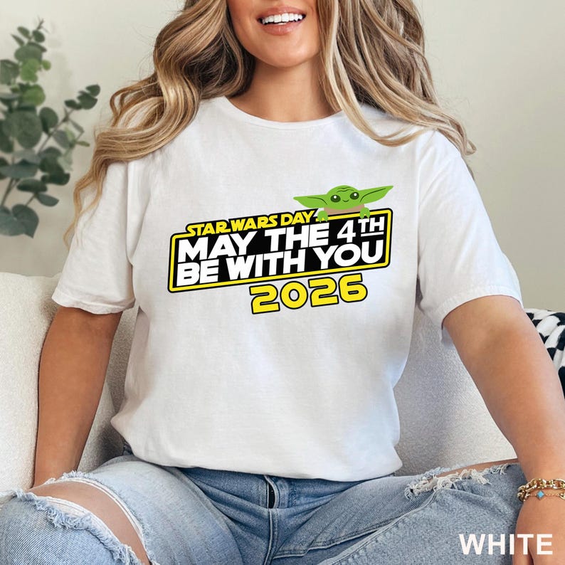 May include: White t-shirt featuring a Star Wars Day graphic. The design includes the text "MAY THE 4TH BE WITH YOU 2026" in yellow and black, with a green Baby Yoda character. The shirt is a short-sleeved crew neck.
