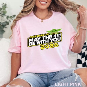 May include: Light pink t-shirt with the text "STAR WARS DAY MAY THE 4TH BE WITH YOU 2026" in yellow and black. The shirt features a green cartoon character. The shirt is made of a soft material.