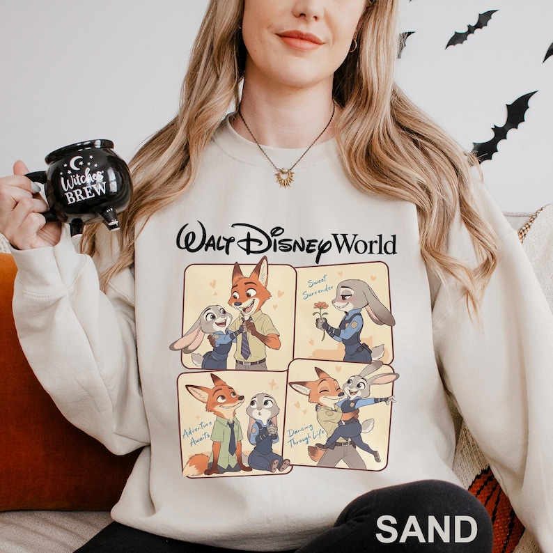 May include: Sand-colored sweatshirt featuring a four-panel graphic of Zootopia characters, with the text "Walt Disney World" above the graphic. The bottom right corner of the sweatshirt has the word "SAND".