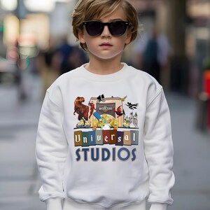 May include: White sweatshirt featuring a colorful graphic of Universal Studios characters, including a T-Rex, The Simpsons, and Spider-Man. The words "Universal Studios" are printed in a block letter design. The sweatshirt is worn by a person wearing sunglasses.