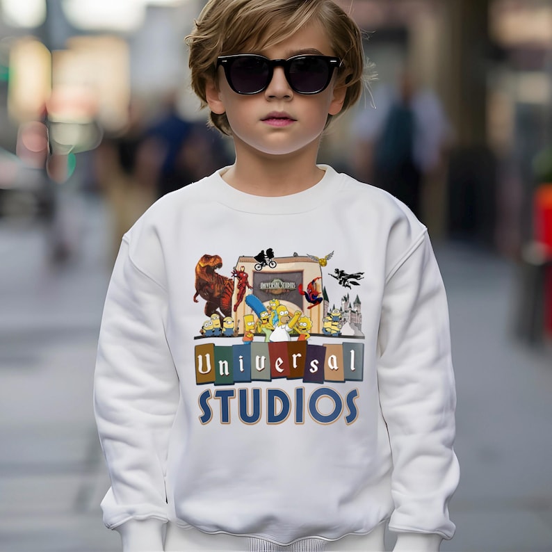 May include: White sweatshirt featuring a colorful graphic of Universal Studios characters, including a T-Rex, The Simpsons, and Spider-Man. The words "Universal Studios" are printed in a block letter design. The sweatshirt is worn by a person wearing sunglasses.
