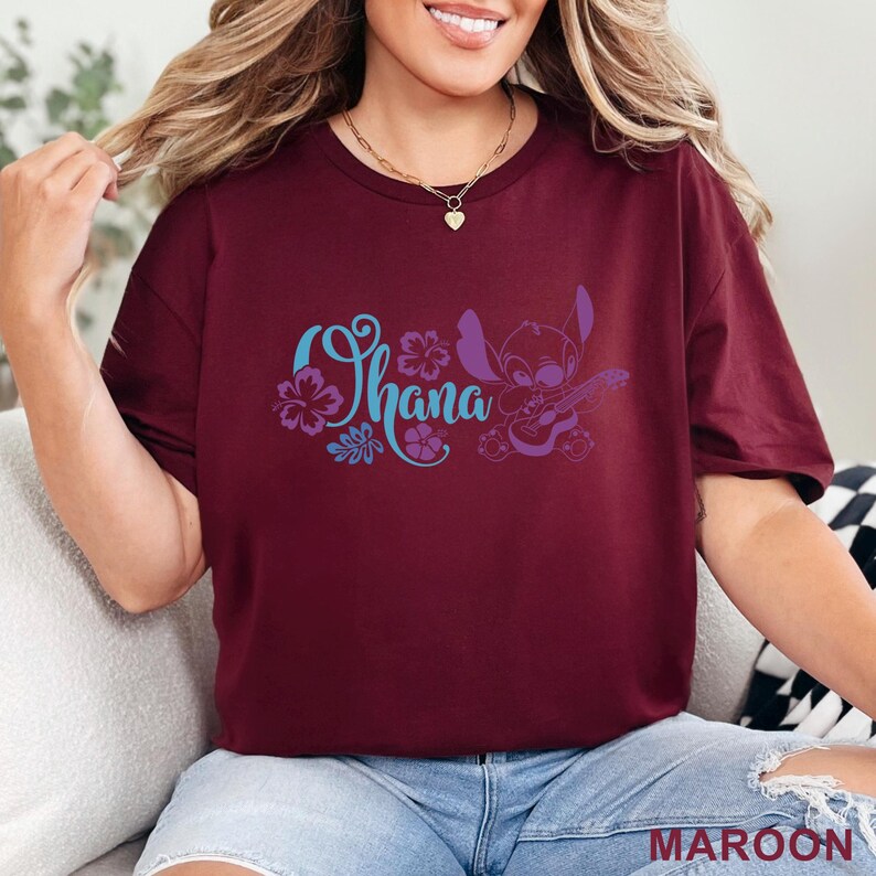 Op de afbeelding: Een bordeauxrood t-shirt met korte mouwen en een grafische print in lichtblauw en paars. Het ontwerp bevat het woord Ohana in een gestileerd cursief lettertype, omringd door hibiscusbloemen en een stripfiguur met een kleine gitaar.