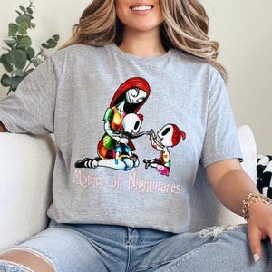 Puede incluir: Camiseta gris con una ilustraci&oacute;n colorida de un personaje con cabello rojo y un vestido de retazos, abrazando a dos figuras de esqueleto. El texto "Mother of Nightmares" est&aacute; impreso en rosa debajo.