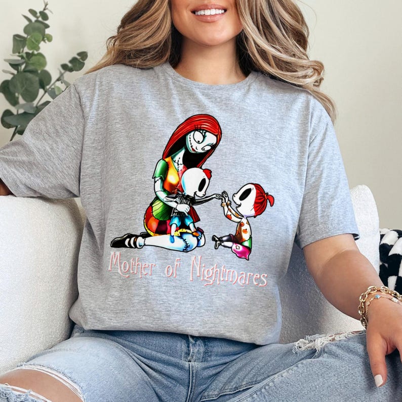Puede incluir: Camiseta gris con una ilustraci&oacute;n colorida de un personaje con cabello rojo y un vestido de retazos, abrazando a dos figuras de esqueleto. El texto "Mother of Nightmares" est&aacute; impreso en rosa debajo.
