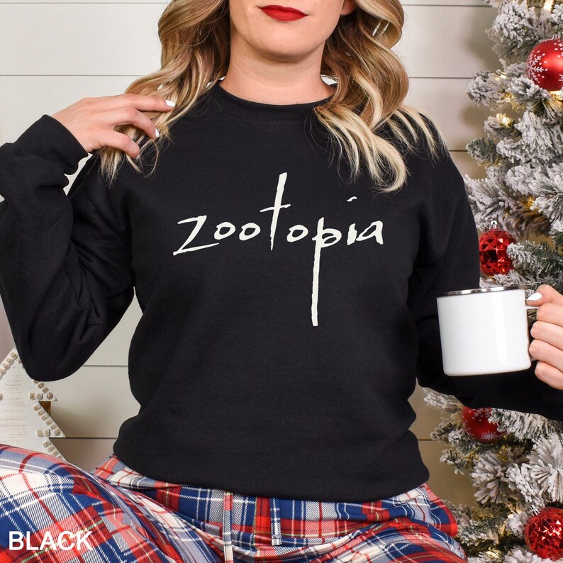 May include: Black crew neck sweatshirt with the word "Zootopia" in white script. The person is holding a white mug with a silver rim. The bottom left corner of the image has the word "BLACK".