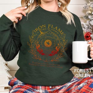 May include: Forest green sweatshirt with the text "IRON FLAME BURN IT DOWN" in gold and red. The design includes a circular emblem with a bird and flames, surrounded by leaves. The bottom of the sweatshirt has a quote.
