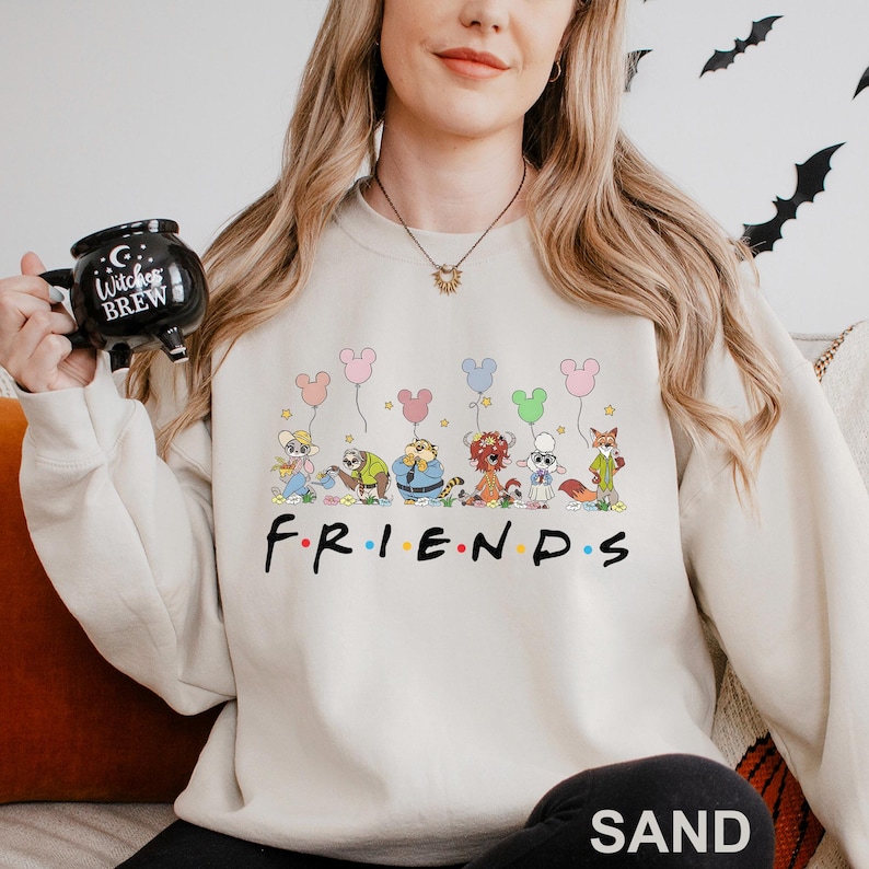 May include: Sand-colored sweatshirt featuring a cartoon graphic of Disney characters holding Mickey Mouse-shaped balloons. The word "FRIENDS" is printed below the characters in a black font. The sweatshirt is a crew neck style.