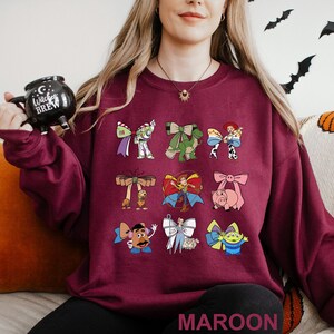 May include: Maroon sweatshirt featuring colorful cartoon character bows. The bows include Buzz Lightyear, Rex, Jessie, Woody, Hamm, Mr. Potato Head, Bo Peep, and a Little Green Alien. The word "MAROON" is printed at the bottom.