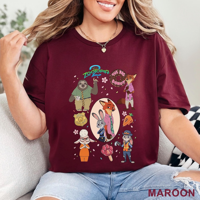 K&ouml;nnte beinhalten: Ein kastanienbraunes Kurzarm-T-Shirt mit einem Zootopia 2-Grafikdruck. Das Design zeigt Charaktere wie Judy Hopps, Nick Wilde und Flash sowie Symbole wie einen Donut, eine Karotte, eine Polizeimarke und ein pfotenf&ouml;rmiges Eis am Stiel.