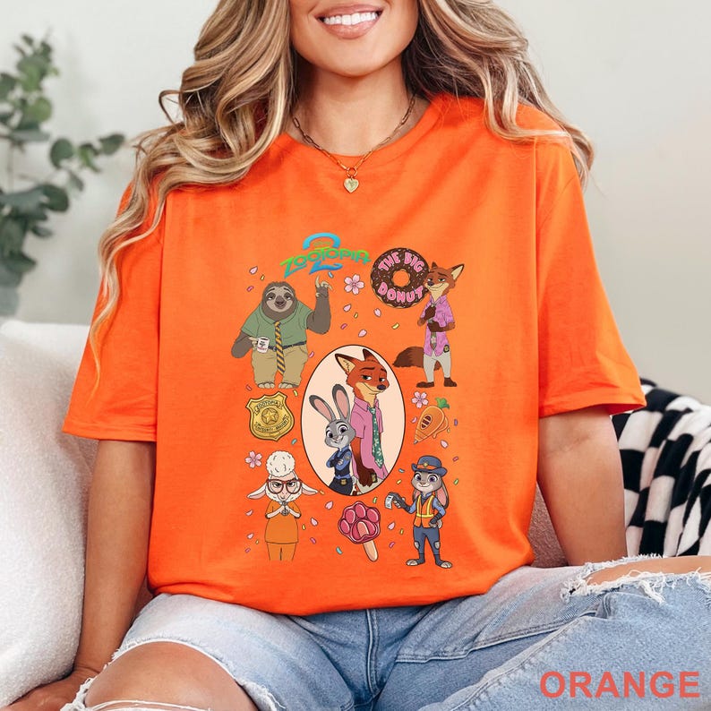 K&ouml;nnte beinhalten: Ein orangefarbenes T-Shirt mit kurzen &Auml;rmeln und einem Zootopia 2-Grafikdruck. Das Design zeigt Charaktere wie Nick Wilde und Judy Hopps, eine Polizeimarke, einen Donut, eine Karotte und ein pfotenf&ouml;rmiges Eis, umgeben von kleinen rosa Bl&uuml;ten.