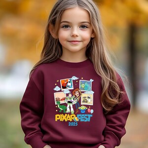 Może przedstawiać: Bordowa bluza z kolorową grafiką postaci z film&oacute;w Pixar i napisem "PIXAR FEST 2025". Projekt zawiera obrazy postaci z r&oacute;żnych film&oacute;w Pixara. Bluza jest noszona przez młodą dziewczynę.