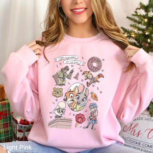 May include: Light pink sweatshirt featuring a colorful graphic of characters and elements from a popular animated movie. The design includes a donut, a police badge, and the text "You can be whatever you wish to be." The sweatshirt is made of a soft material.