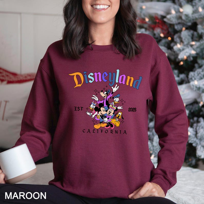 Puede incluir: Sudadera burdeos con el logotipo de Disneyland y un gr&aacute;fico de Mickey Mouse, Minnie Mouse, Donald Duck, Goofy y Pluto. El texto "EST 2025 CALIFORNIA" tambi&eacute;n est&aacute; impreso en la sudadera.