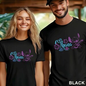 Op de afbeelding: Een zwart t-shirt met korte mouwen en een grafische print van een paars stripfiguur dat ukelele speelt. Het ontwerp bevat het woord Ohana in een lichtblauw lettertype, omringd door paarse en blauwe illustraties van hibiscusbloemen.