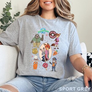 K&ouml;nnte beinhalten: Ein hellgraues T-Shirt mit einem farbenfrohen Zootopia 2 Grafikdruck. Das Design zeigt Charaktere wie Judy Hopps und Nick Wilde, eine Polizeimarke, einen Donut, eine Karotte und ein pfotenf&ouml;rmiges Eis am Stiel in einer Collage-Anordnung.