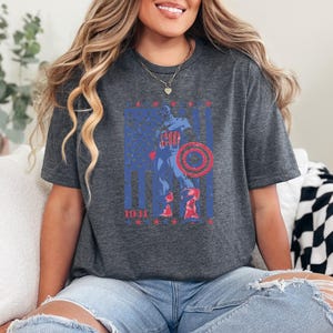 May include: Dark gray t-shirt featuring a distressed graphic of a superhero with a shield, set against a blue and red American flag design. The year "1941" is printed at the bottom. The shirt has a crew neck and short sleeves.