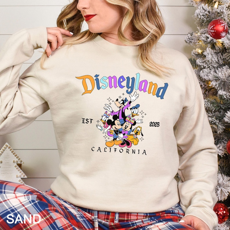 Puede incluir: Una sudadera de color arena con el logotipo de Disneyland en naranja y azul, con un gr&aacute;fico de Mickey Mouse, Minnie Mouse, Donald Duck, Goofy y Pluto. El texto "EST 2025 CALIFORNIA" tambi&eacute;n est&aacute; impreso.
