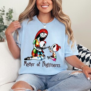 Puede incluir: Camiseta azul claro con un gr&aacute;fico colorido de un personaje con cabello rojo y un vestido de retazos, con dos ni&ntilde;os esqueleto. El texto "Mother of Nightmares" est&aacute; impreso debajo. La camiseta es de material suave.