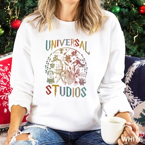 Pode incluir: Camisola branca com um gr&aacute;fico colorido da Universal Studios. O design inclui personagens e atra&ccedil;&otilde;es de parques tem&aacute;ticos, com o texto "UNIVERSAL STUDIOS" em arco acima e abaixo. A camisola &eacute; branca lisa.