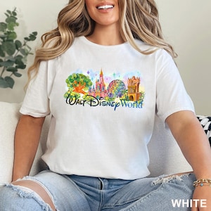 May include: A white short-sleeved t-shirt featuring a colorful watercolor-style graphic of Walt Disney World landmarks, including a tree, a castle, a geodesic dome, and a tower, with the text Walt Disney World printed in a dark blue script font.