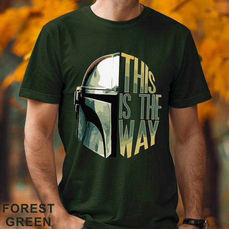 Pu&ograve; includere: Una t-shirt a maniche corte color verde bosco con una stampa grafica di un casco metallico sul lato sinistro e il testo THIS IS THE WAY in lettere maiuscole gialle e verdi sul lato destro, centrato sul petto del capo di abbigliamento.