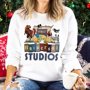 May include: White sweatshirt with a colorful graphic featuring characters from Universal Studios attractions, including dinosaurs, Minions, and The Simpsons. The text "Universal STUDIOS" is displayed in blue and brown letters.