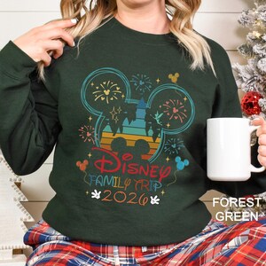 May include: Forest green sweatshirt with a colorful Disney Family Trip 2026 graphic. The design features Mickey Mouse ears, fireworks, and a castle silhouette in shades of blue, orange, and red. A white mug is held in the hand.