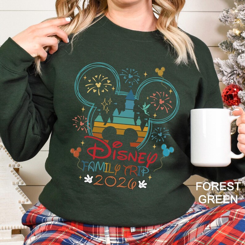 May include: Forest green sweatshirt with a colorful Disney Family Trip 2026 graphic. The design features Mickey Mouse ears, fireworks, and a castle silhouette in shades of blue, orange, and red. A white mug is held in the hand.