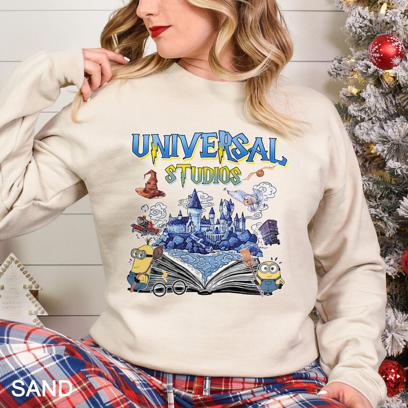 Op de afbeelding: Zandkleurige sweatshirt met een kleurrijke afbeelding van het Universal Studios-logo en themaparkelementen. Het ontwerp bevat een tovenaars hoed, een kasteel en Minions. De tekst "UNIVERSAL STUDIOS" is in blauw en geel.