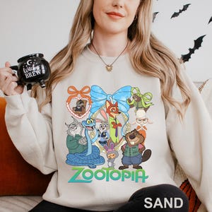 May include: Sand-coloured sweatshirt with a colourful graphic of characters from the animated film "Zootopia." The design includes a blue bow, a heart-shaped frame, and the word "Zootopia" in green. The word "SAND" is printed at the bottom.