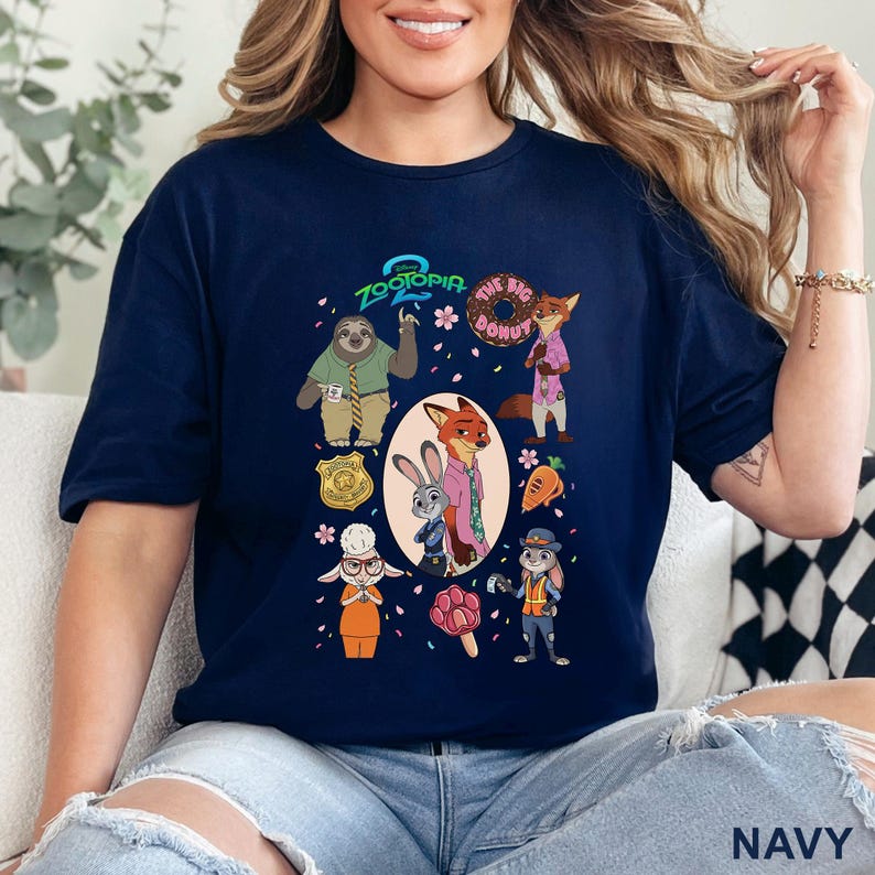 K&ouml;nnte beinhalten: Ein marineblaues T-Shirt mit einem Zootopia-Grafikdruck. Das Design zeigt die Figuren Flash, Nick Wilde, Judy Hopps und Bellwether sowie Symbole wie einen Donut, eine Karotte, ein pfotenf&ouml;rmiges Eis am Stiel und ein Polizeimarke, umgeben von Bl&uuml;ten.